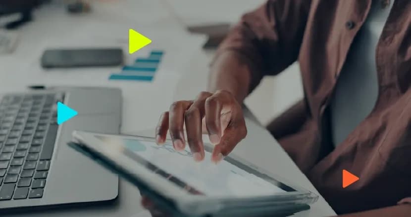 Profissional analisa gráficos financeiros na tela de um tablet. Sobre a mesa de trabalho há também um notebook aberto, papéis impressos com relatórios e um celular.
