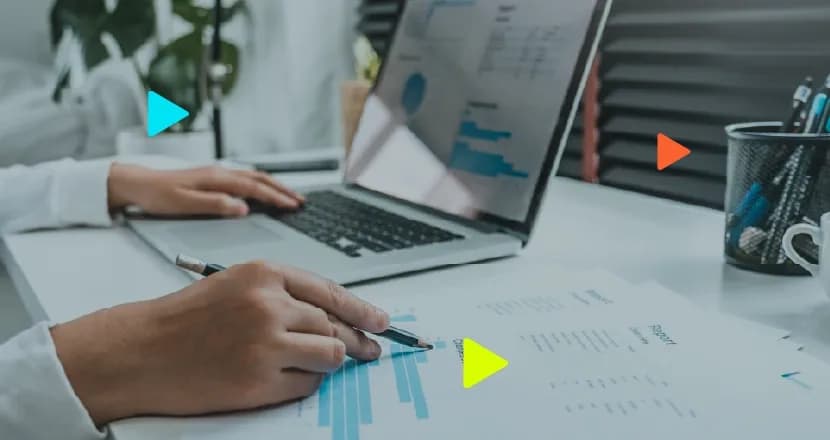Pessoa analisa relatório com gráficos usando lápis, com notebook aberto exibindo dados na mesa de trabalho.
