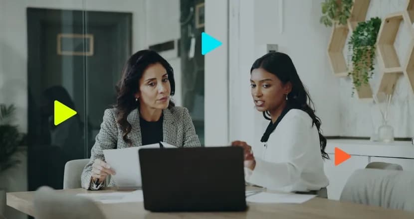 Duas mulheres sentadas à mesa analisando um notebook; uma delas segura documentos impressos. Ícones coloridos de 'play' aparecem sobrepostos na imagem.