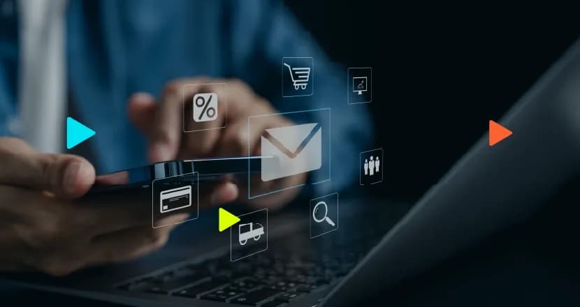 Mãos seguram um smartphone sobre o teclado de um notebook em um ambiente escuro. Da tela do celular surgem ícones digitais flutuantes brancos, como um envelope de email, cartão de crédito e carrinho de compras.