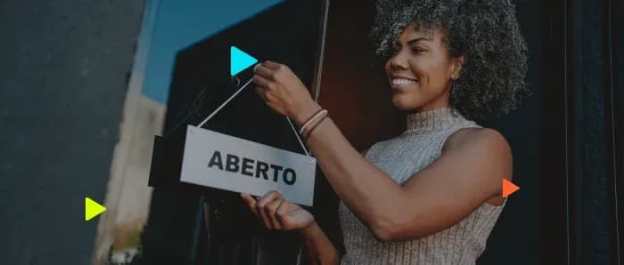 Empreendedora sorridente pendura placa de ABERTO na porta, ilustrando a abertura de CNPJ MEI.