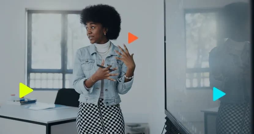 Mulher negra com cabelo afro, vestindo jaqueta jeans e saia xadrez, gesticulando com as mãos e segurando uma caneta durante uma apresentação ao lado de um quadro.